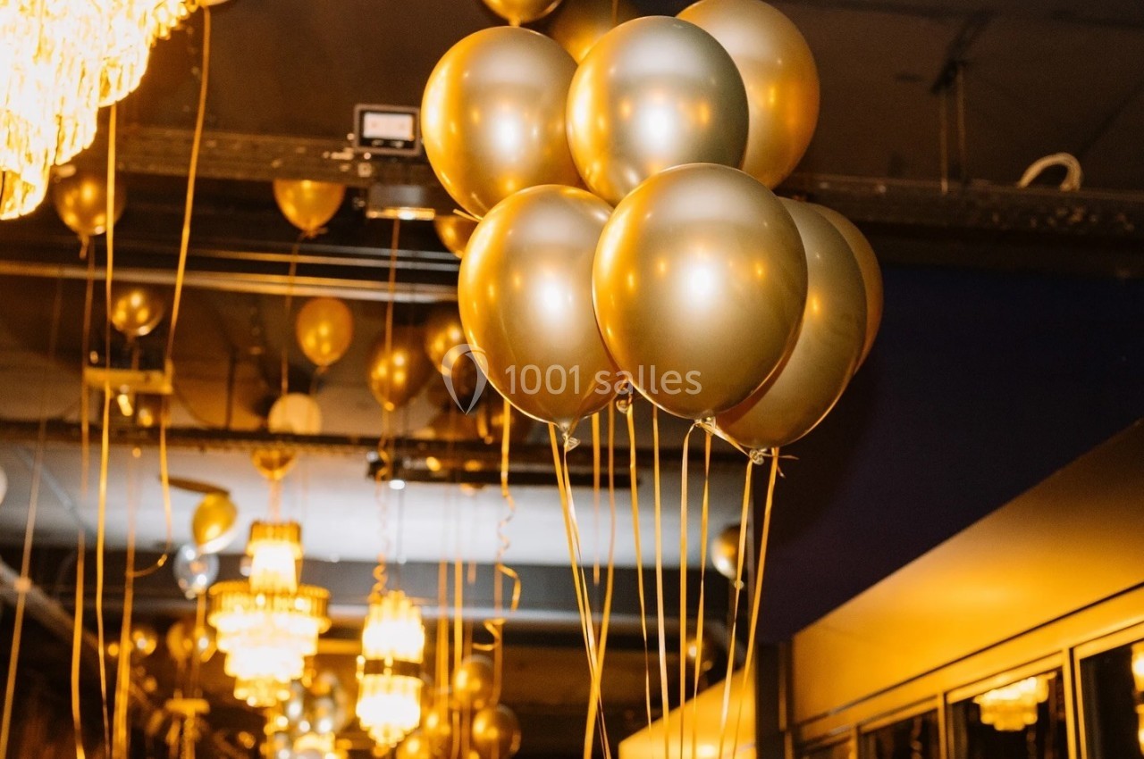 Des ballons dorés flottent sous un plafond éclairé par des lustres dans une salle décorée pour un événement.