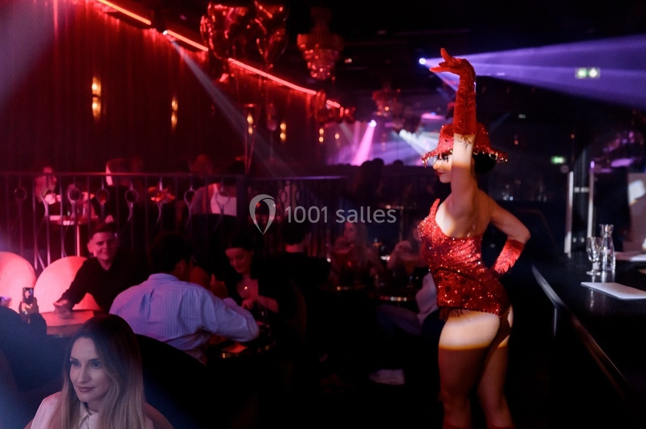 Danseuse en tenue rouge scintillante sur scène dans un cabaret, éclairé par des lumières rouges et violettes.