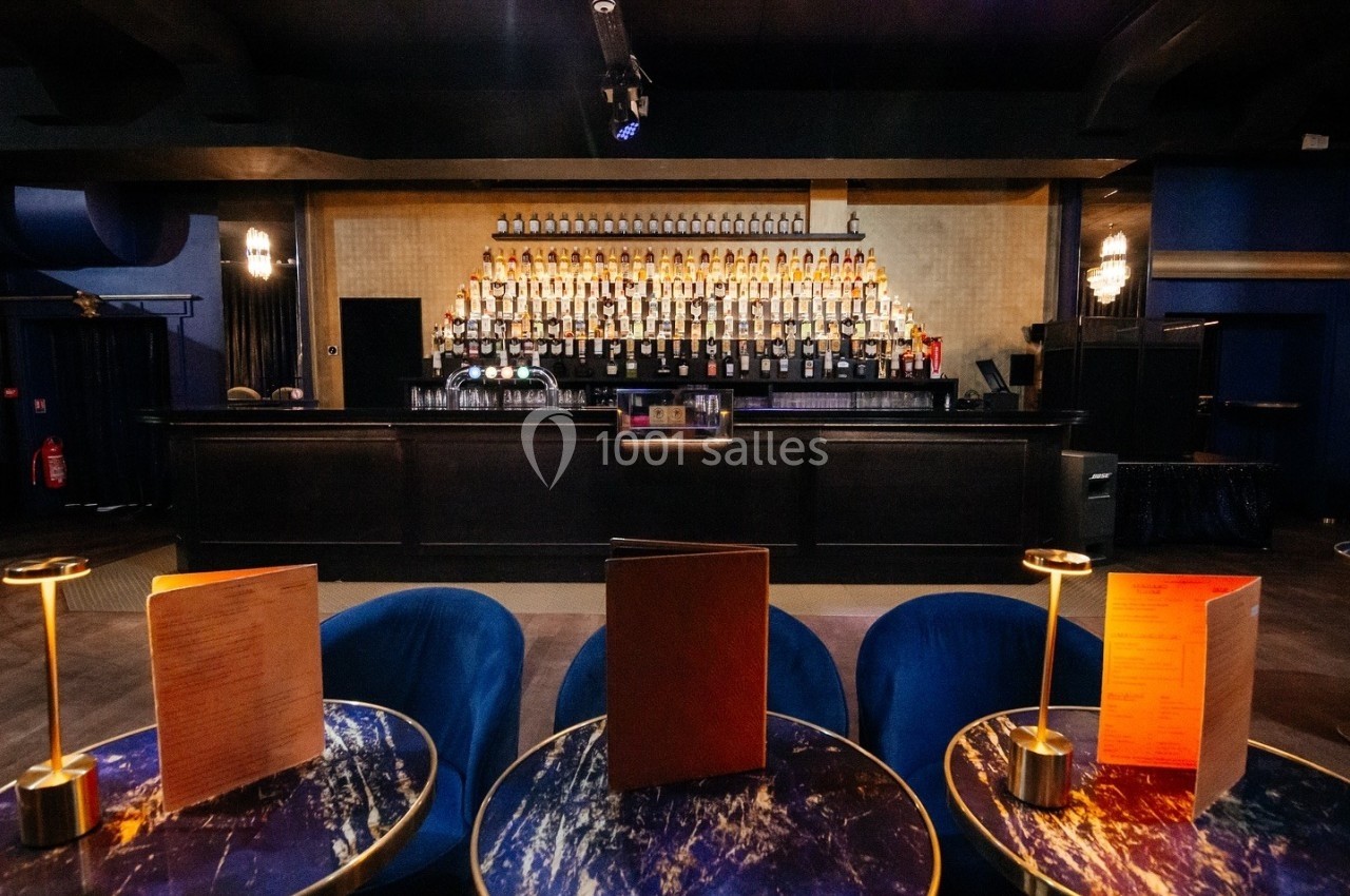 Bar élégant avec comptoir en bois sombre, étagères remplies de bouteilles et tables avec lampes et menus au premier plan.