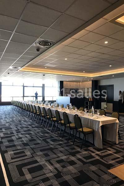 Salle de réception lumineuse avec une longue table dressée pour un repas, chaises alignées et bar en arrière-plan.