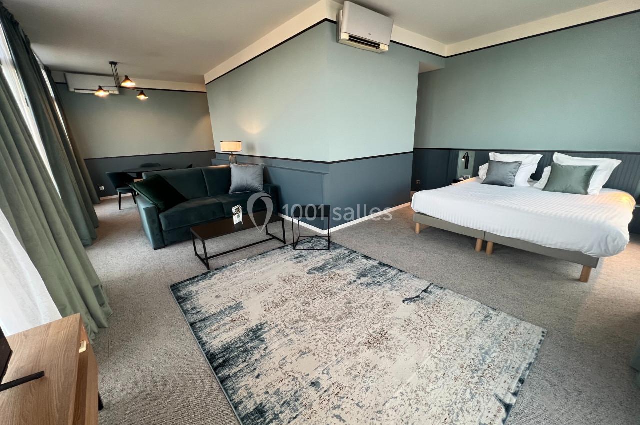 Chambre spacieuse avec lit double, canapé, tapis moderne, éclairage doux et murs aux tons verts et gris.