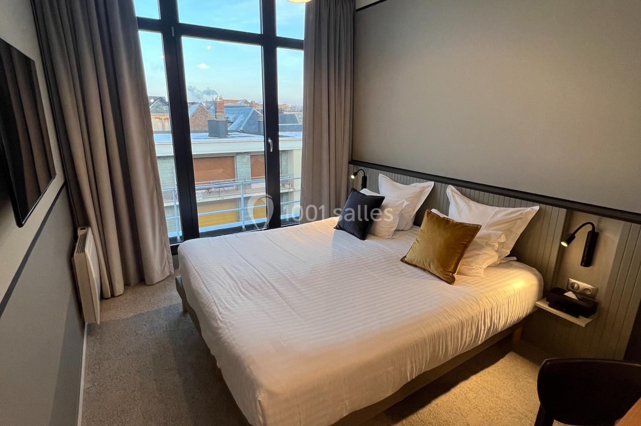 Chambre moderne avec lit double, grandes fenêtres donnant sur des bâtiments, éclairage suspendu et décor sobre.