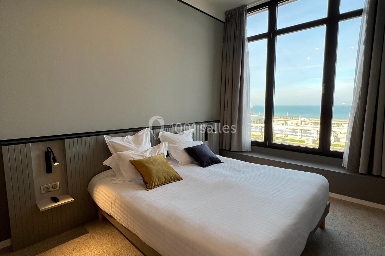 Chambre lumineuse avec un lit double, des coussins colorés, une grande fenêtre donnant sur une vue maritime.