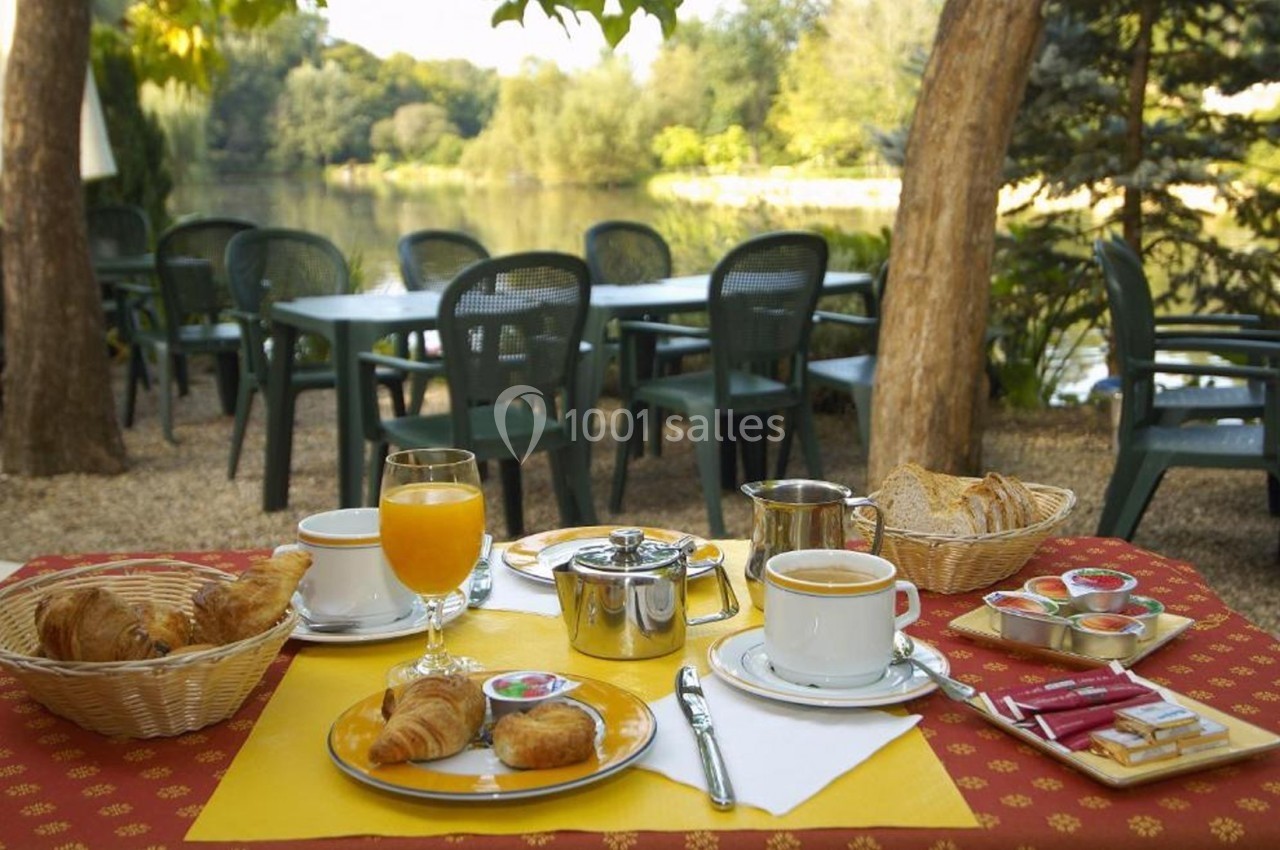 Petit-déjeuner servi en extérieur avec croissants, café, jus d'orange et vue sur un lac entouré de verdure.