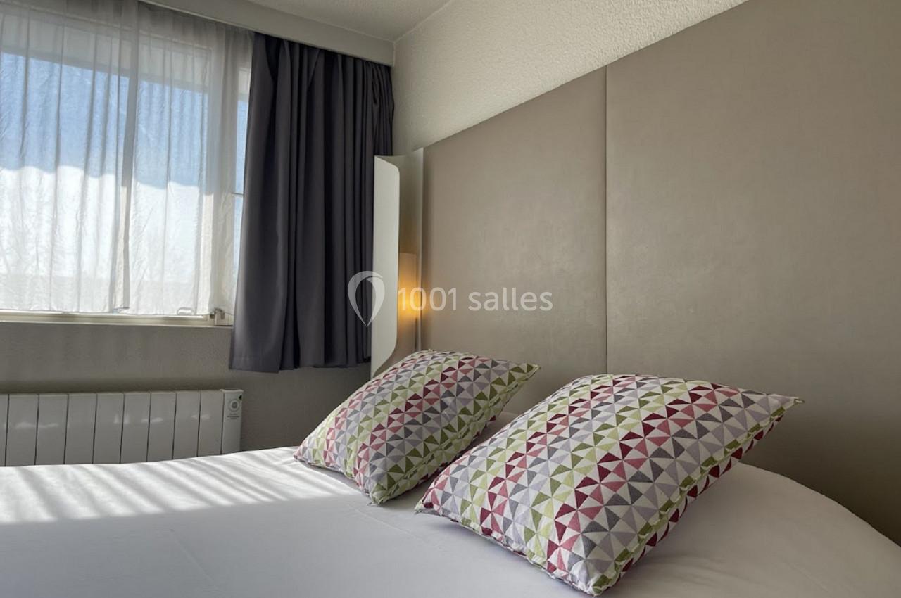 Lit double avec oreillers à motifs géométriques colorés, près d'une fenêtre avec rideaux laissant passer la lumière.