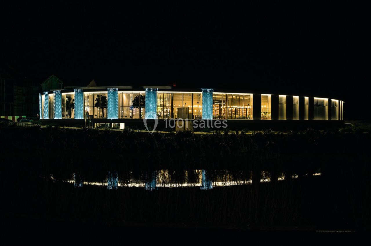 Bâtiment moderne éclairé la nuit, avec des colonnes lumineuses et son reflet visible dans l'eau au premier plan.