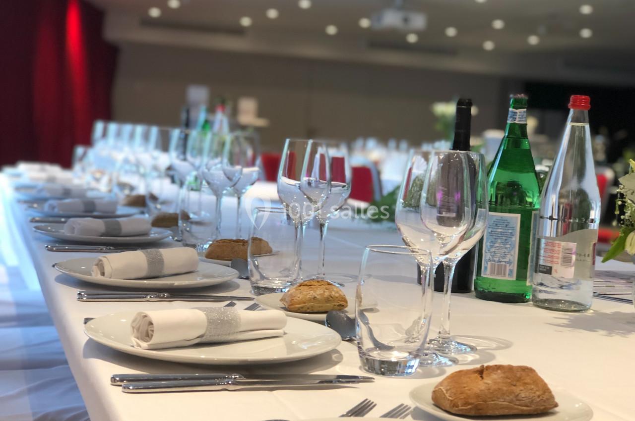 Table dressée avec nappes blanches, verres, assiettes, serviettes pliées, bouteilles d'eau et pain, dans une salle éclairée.
