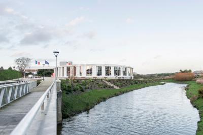 Bâtiment moderne en bord de rivière, entouré de verdure, avec un pont piétonnier au premier plan.
