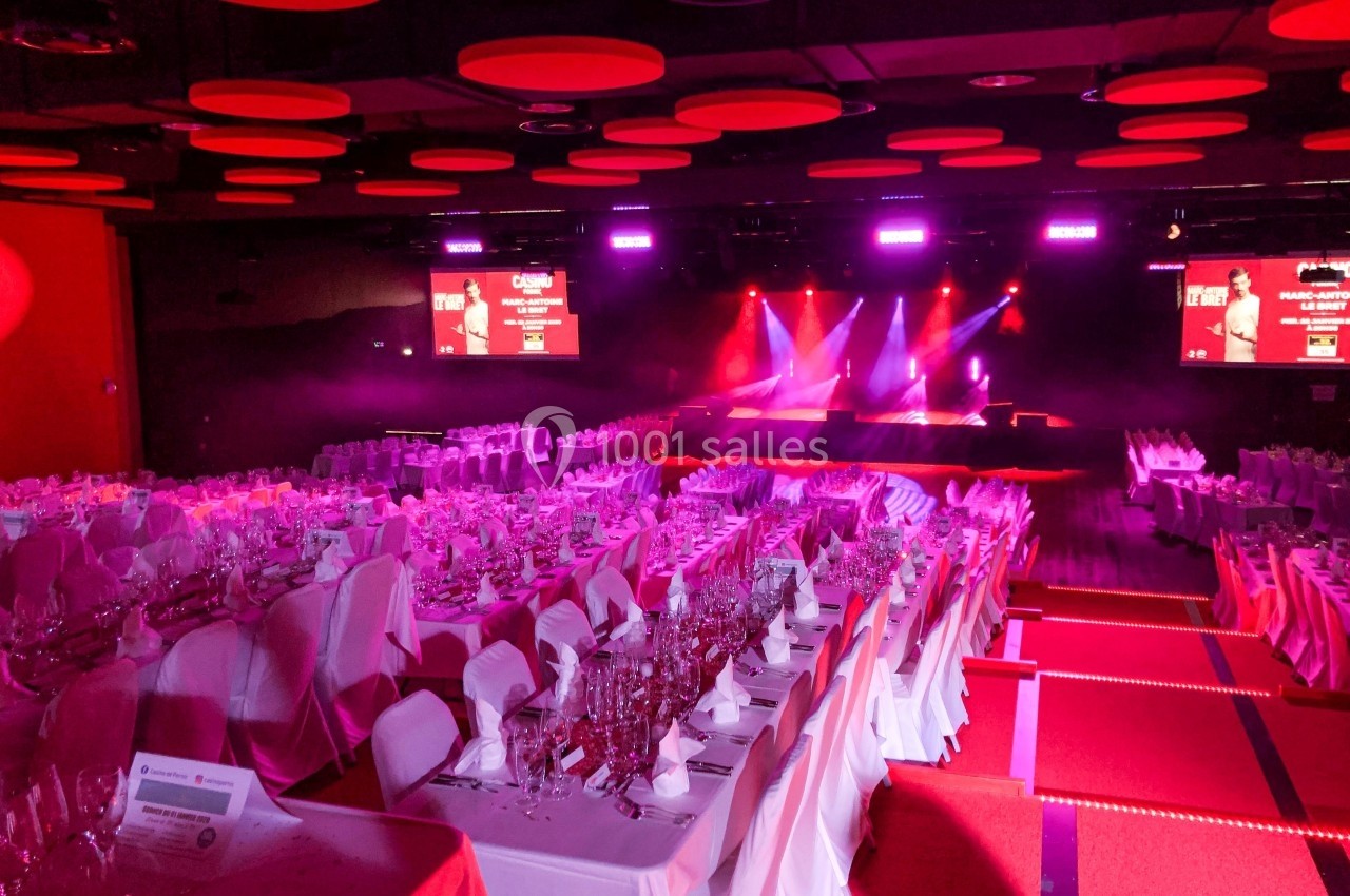 Salle de réception avec tables dressées, éclairage rose et scène illuminée en arrière-plan.
