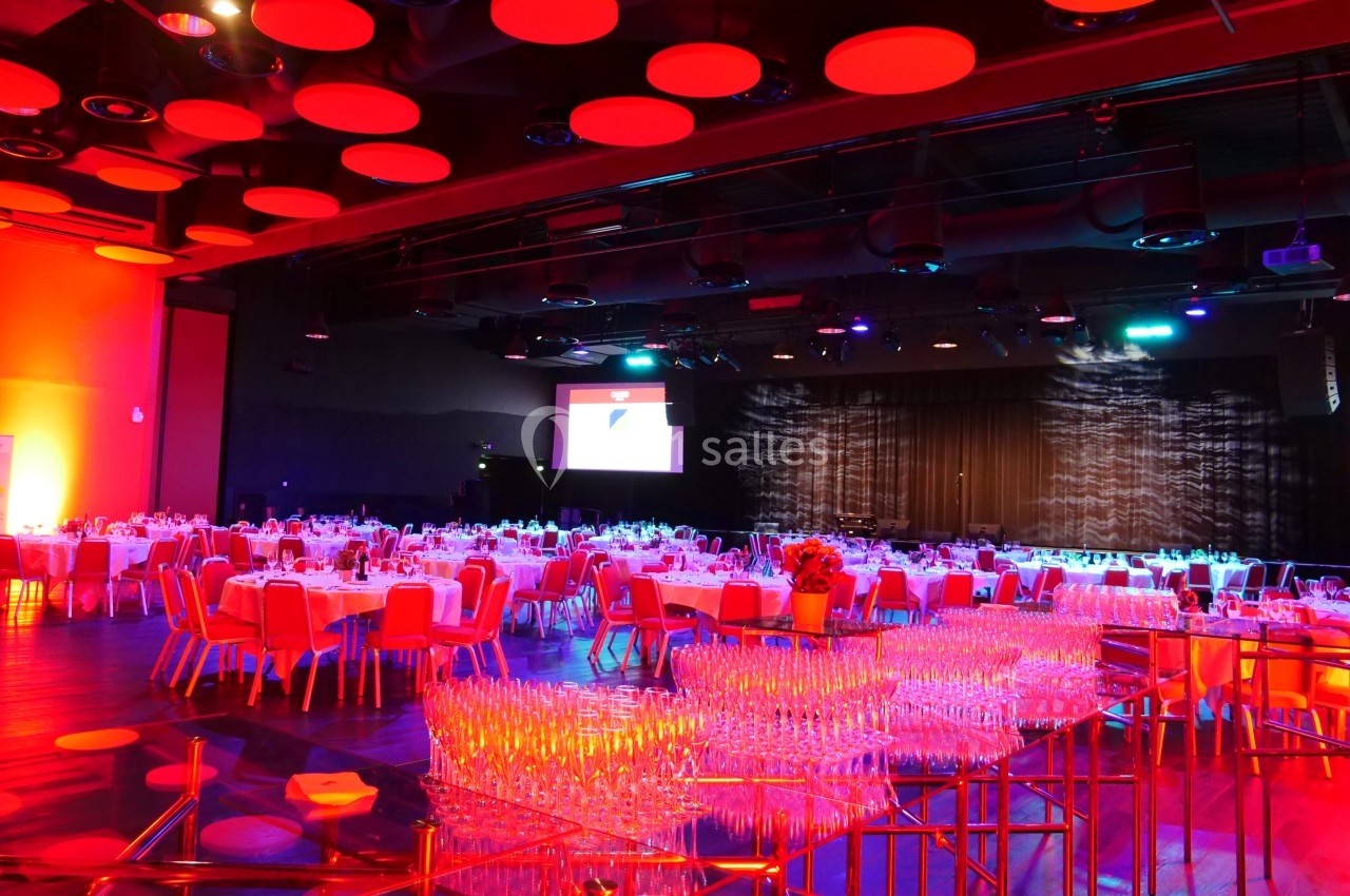 Salle de réception illuminée en rouge avec des tables dressées et un écran de projection en arrière-plan.