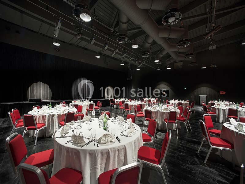 Salle de réception avec tables rondes dressées, nappes blanches et chaises rouges, éclairée par des spots au plafond.