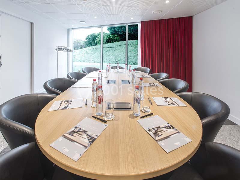 Salle de réunion lumineuse avec une grande table ovale, chaises noires, documents et bouteilles d'eau disposés.