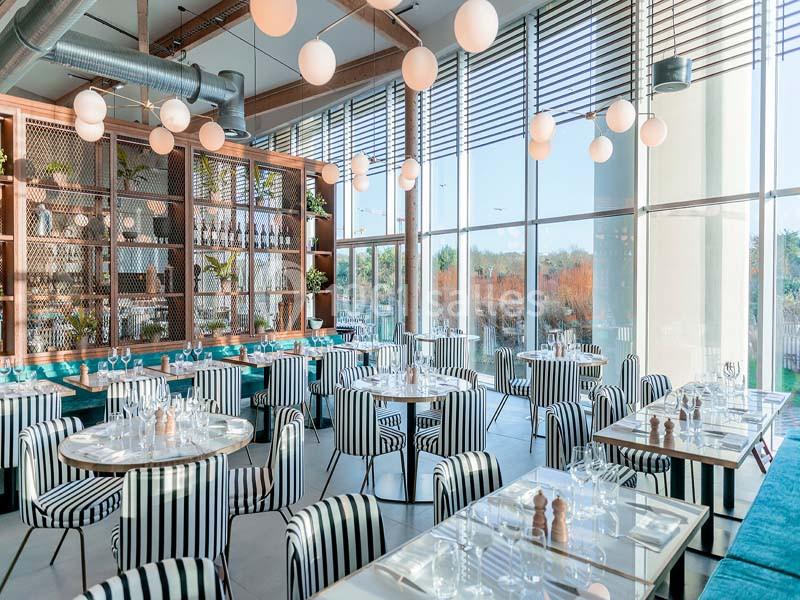 Salle de restaurant lumineuse avec des tables dressées, des chaises à rayures noires et blanches, et de grandes baies…