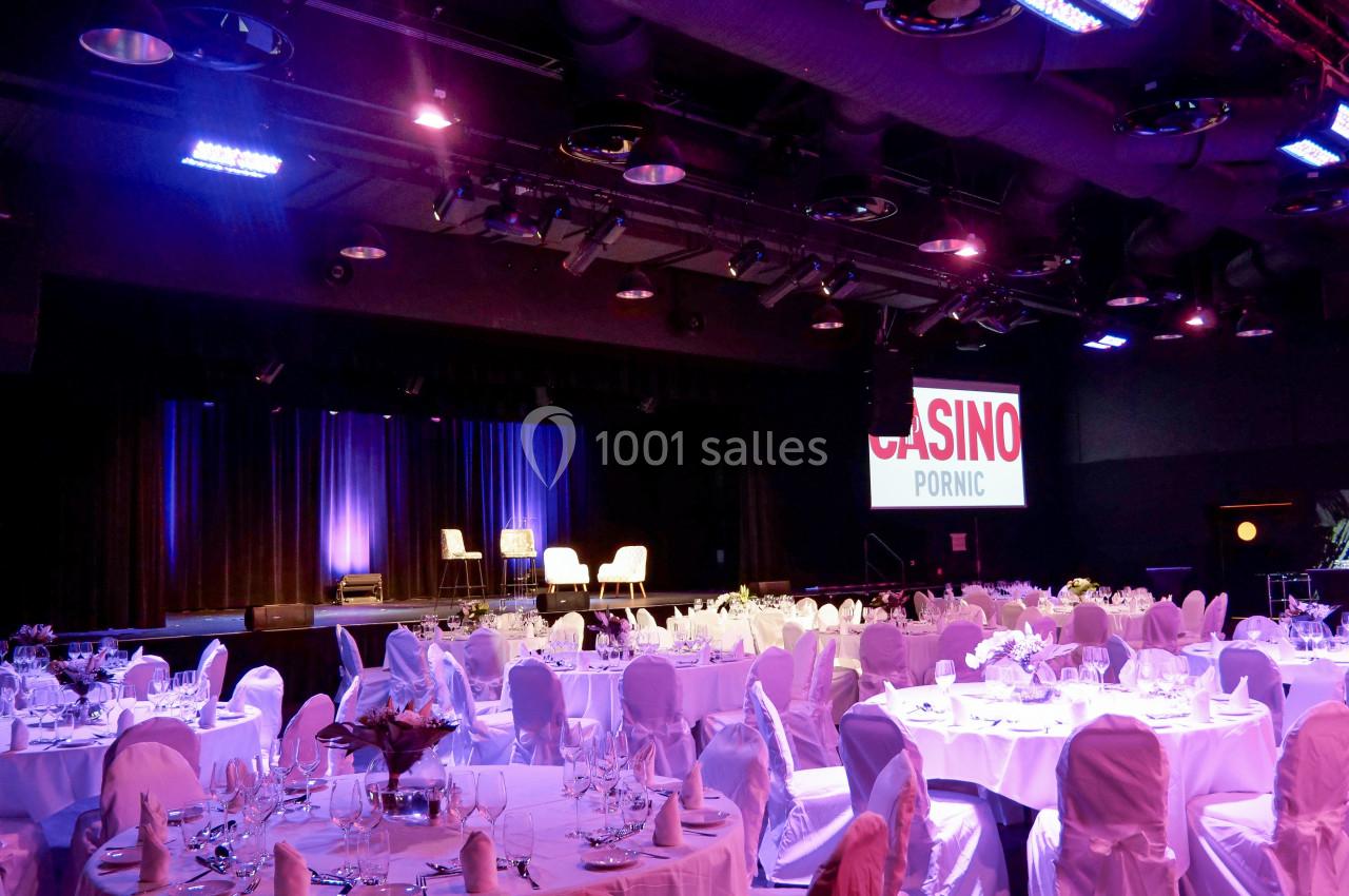Salle de réception avec tables dressées, éclairage tamisé et scène équipée de fauteuils, dans un casino.