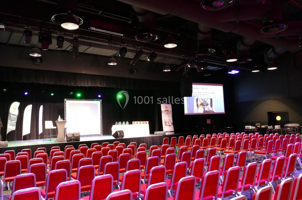 Salle de conférence avec rangées de chaises rouges, une scène éclairée et un écran de projection allumé.