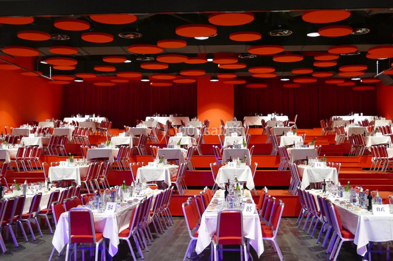 Salle de réception avec des tables dressées, nappes blanches et chaises rouges, éclairage chaleureux.
