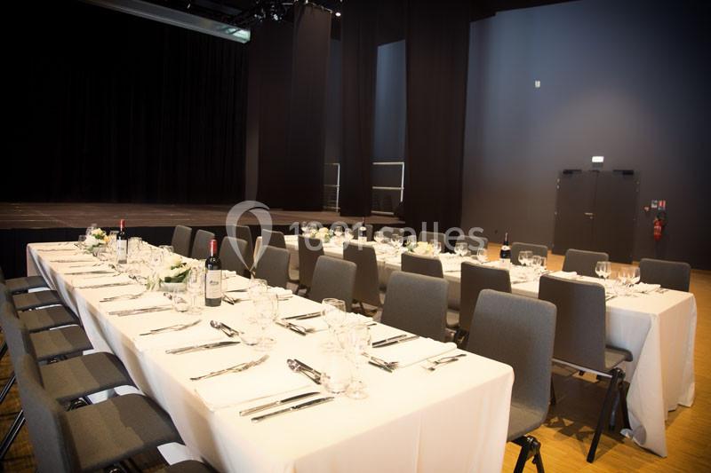 Salle de réception avec tables dressées, nappes blanches, couverts, verres et bouteilles, devant une scène sombre.