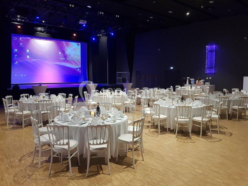Salle de réception avec tables rondes dressées, chaises blanches et grand écran de projection allumé en arrière-plan.