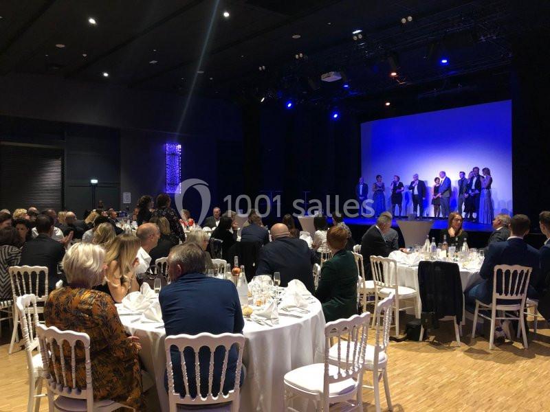 Salle de réception avec des invités assis à des tables rondes, face à une scène où se tiennent plusieurs personnes.