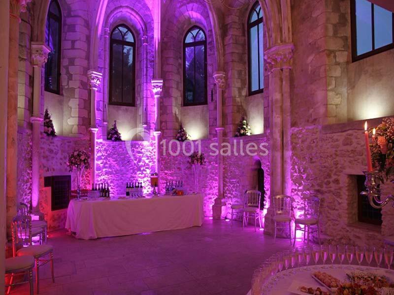 Salle en pierre avec éclairage rose, grandes fenêtres gothiques, tables dressées et décorations élégantes.