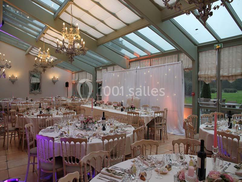Salle de réception élégante avec tables dressées, chaises en bois clair, lustres et grandes baies vitrées donnant sur un…