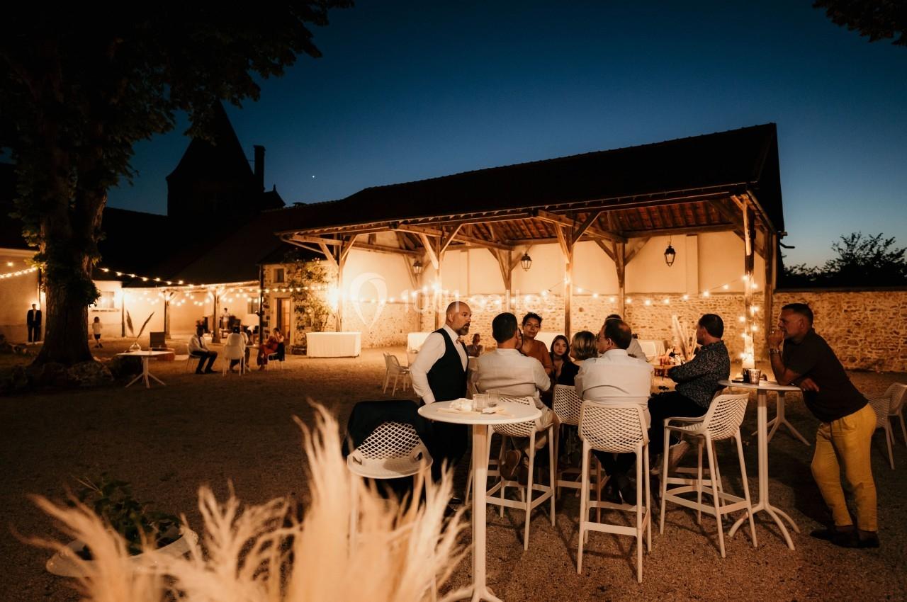 Groupe de personnes discutant autour de tables hautes dans une cour éclairée par des guirlandes lumineuses au crépuscule.