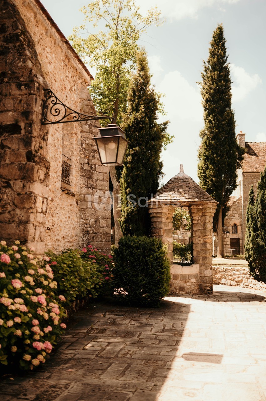 Allée pavée bordée de fleurs et de cyprès, avec un puits en pierre et des bâtiments en pierre sous un ciel ensoleillé.