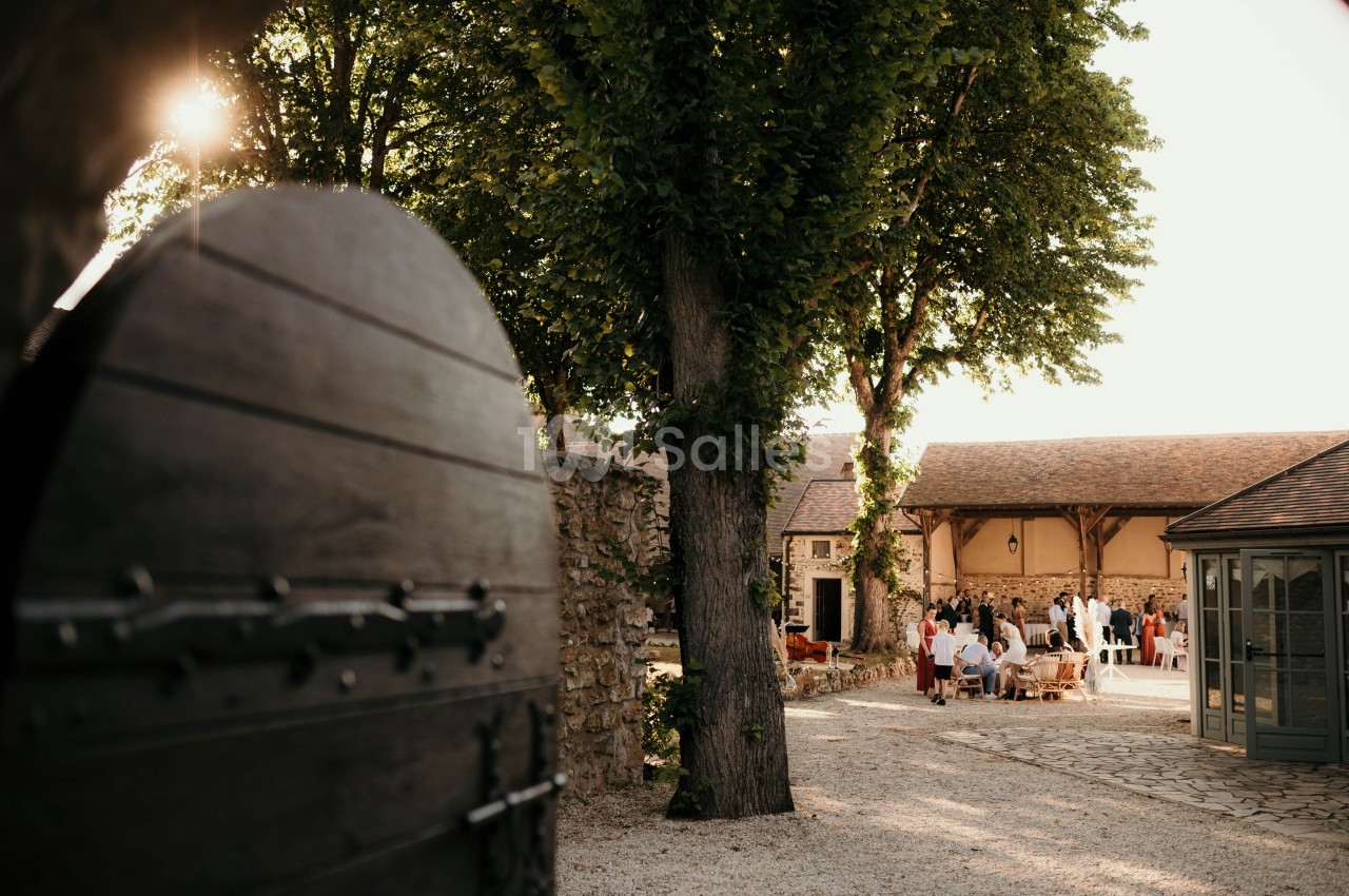 Cour intérieure ensoleillée avec des invités rassemblés près d'un bâtiment en pierre, vue depuis une porte en bois ouverte.
