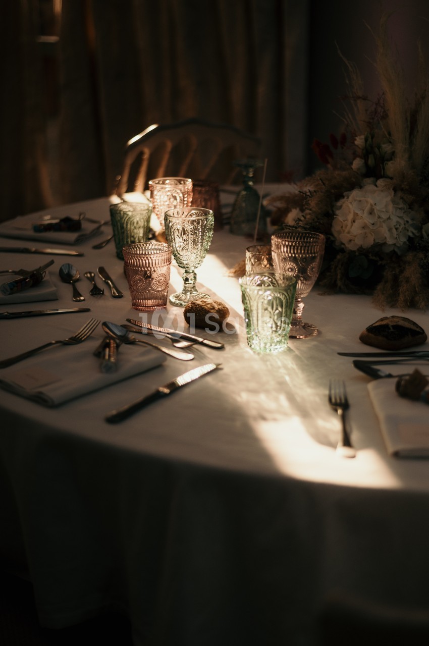 Table dressée avec nappes blanches, verres colorés, couverts et décoration florale dans une lumière tamisée.