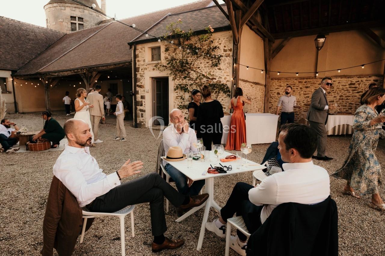 Des personnes discutent autour d'une table en extérieur dans une cour pavée, avec des bâtiments en pierre en arrière-plan.