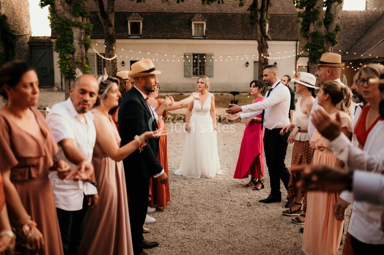 Des invités forment une haie d'honneur pour une mariée en robe blanche dans une cour décorée de guirlandes lumineuses.