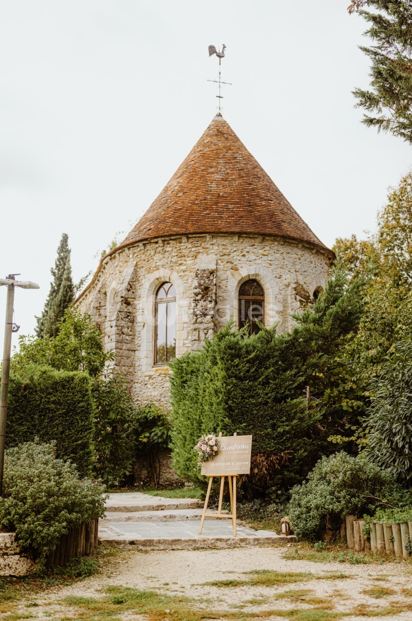 Petite chapelle en pierre avec toit en tuiles, entourée de verdure, et un panneau décoratif sur un chevalet devant.