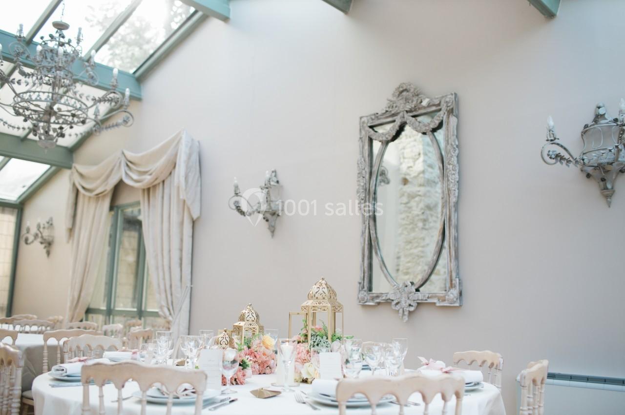 Salle lumineuse avec table décorée de fleurs et lanternes dorées, entourée de chaises et ornée de miroirs et lustres.
