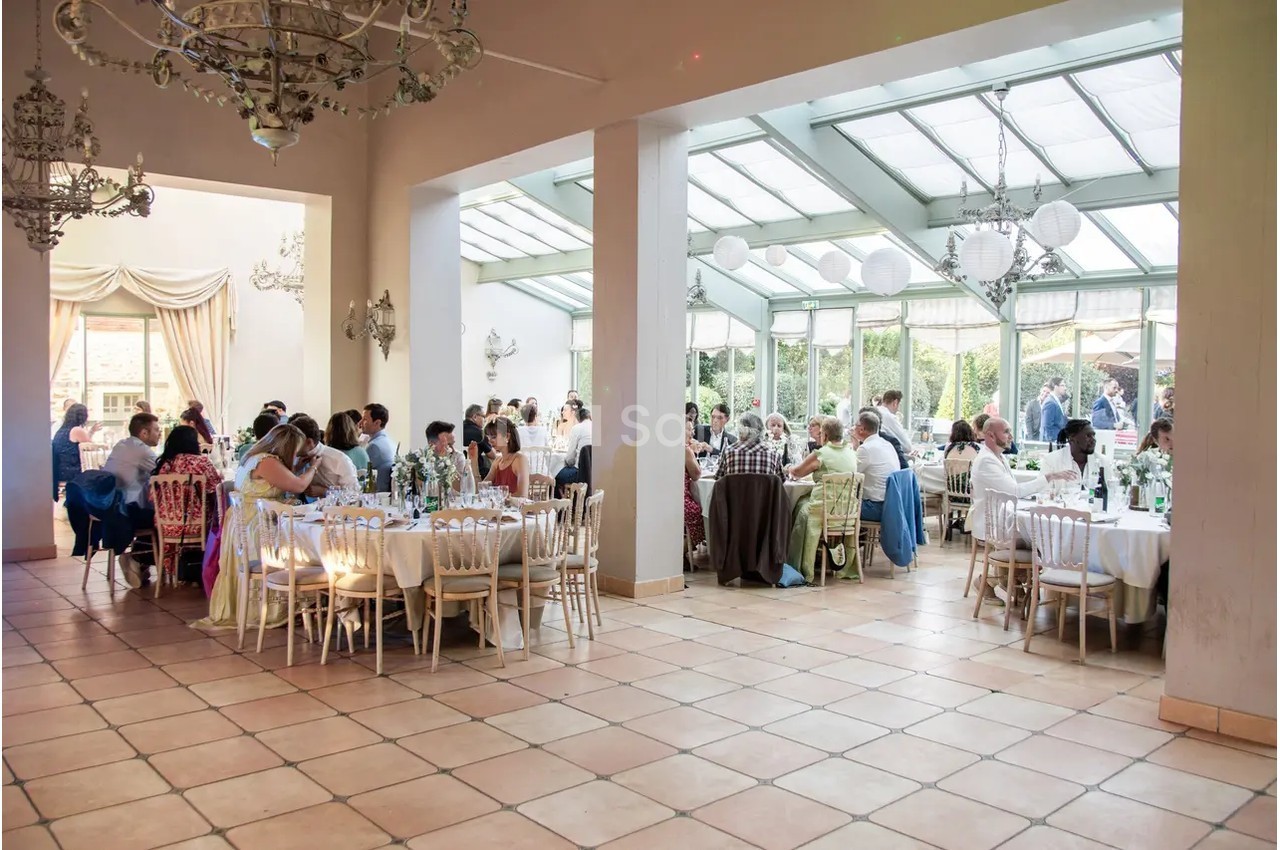 Salle lumineuse avec de grandes baies vitrées, décorée pour un événement, où des invités sont assis à des tables rondes.