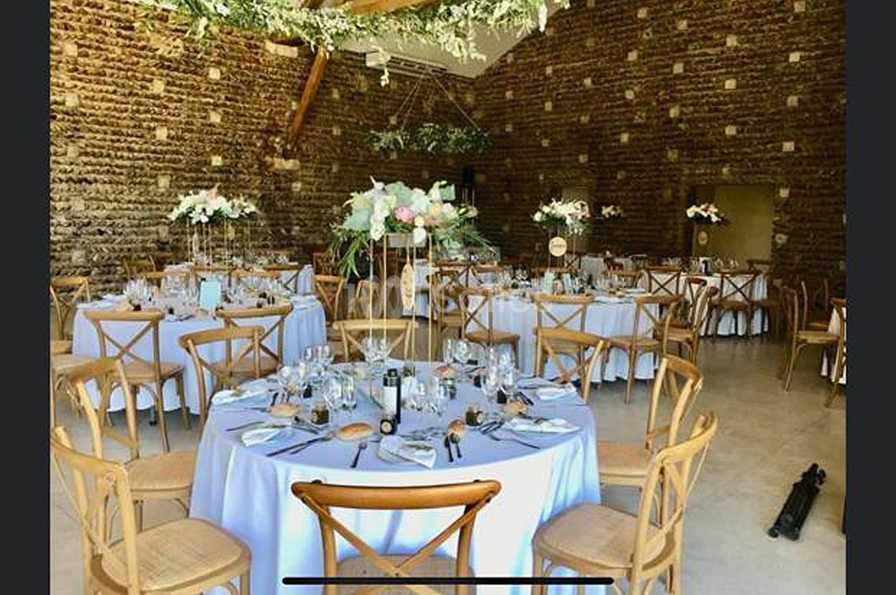 Salle de réception décorée avec des tables rondes dressées, nappes blanches, chaises en bois et arrangements floraux.