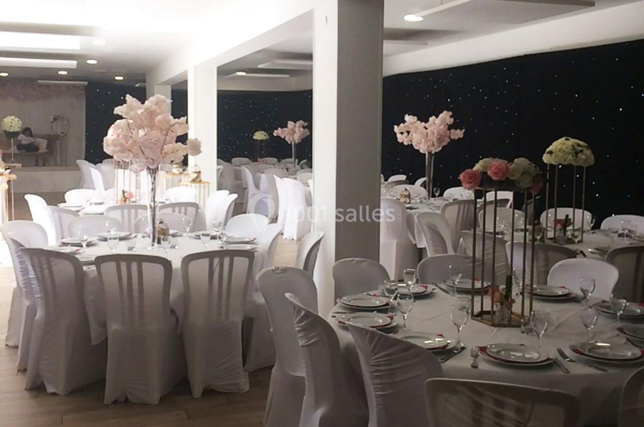 Salle de réception décorée avec des tables rondes, nappes blanches, chaises assorties et centres de table fleuris.