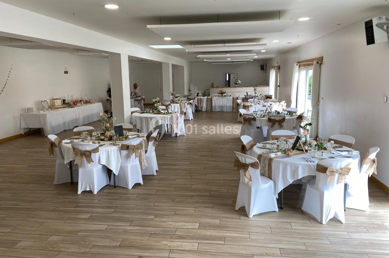 Salle de réception décorée pour un événement, avec des tables rondes dressées et des chaises ornées de nœuds beiges.