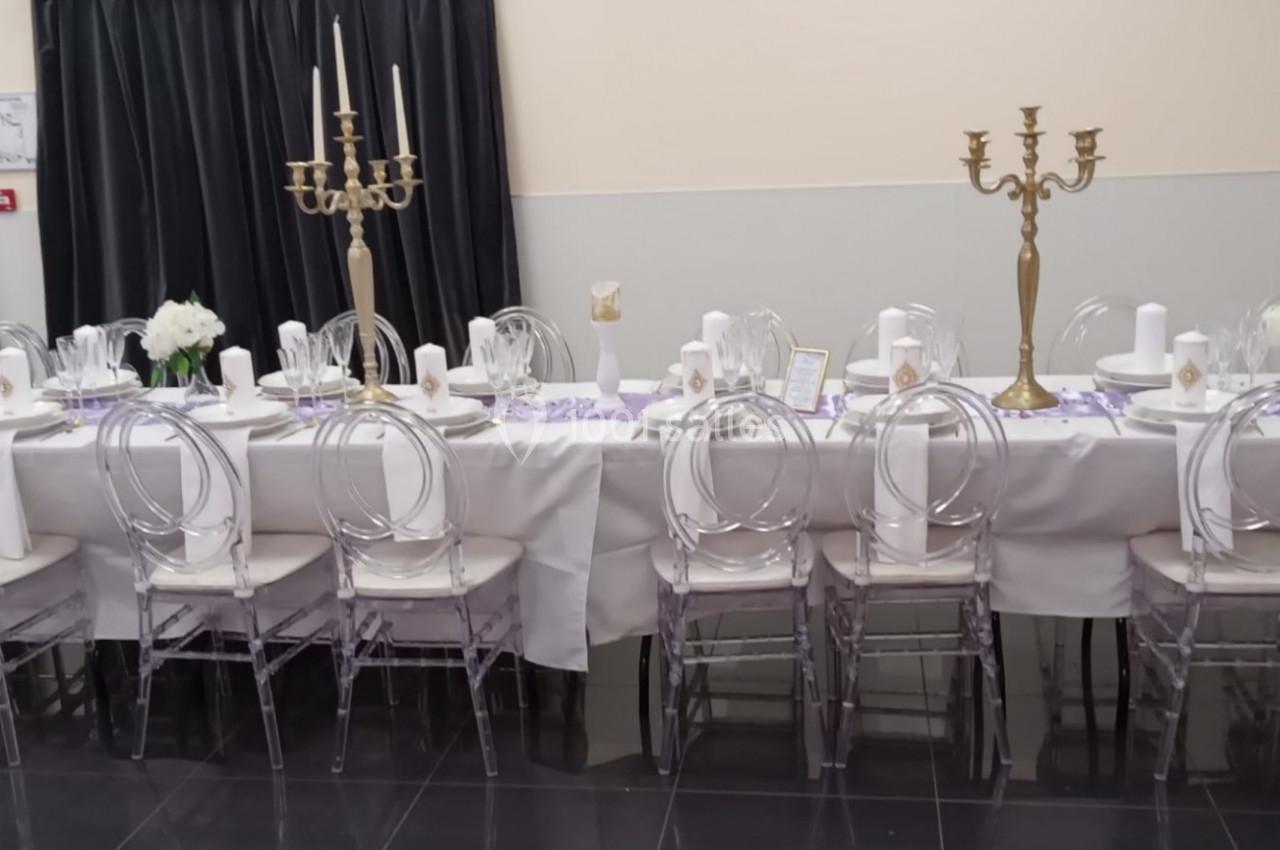 Table dressée pour un événement avec nappes blanches, chaises transparentes, chandeliers dorés et décorations florales.