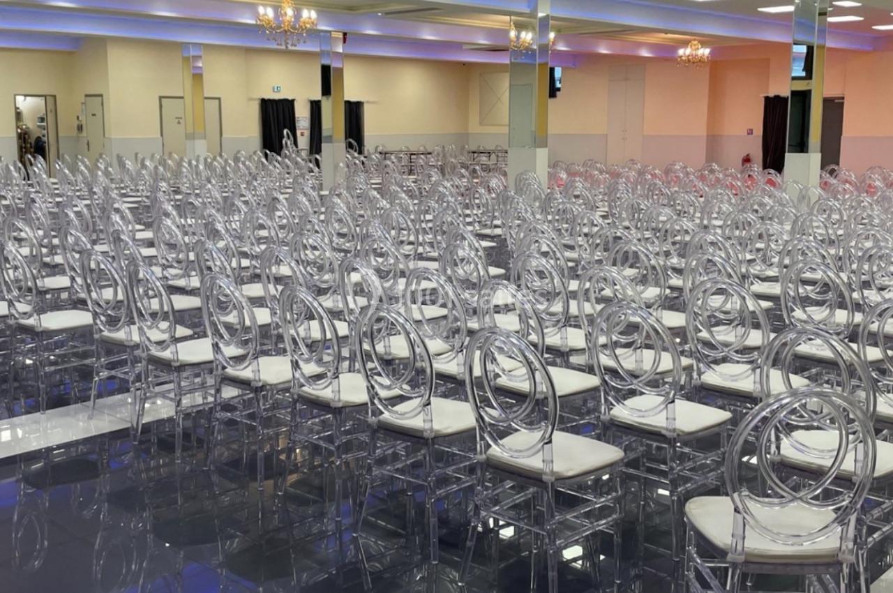 Salle de réception avec de nombreuses chaises transparentes alignées sur un sol brillant, éclairée par des lustres.