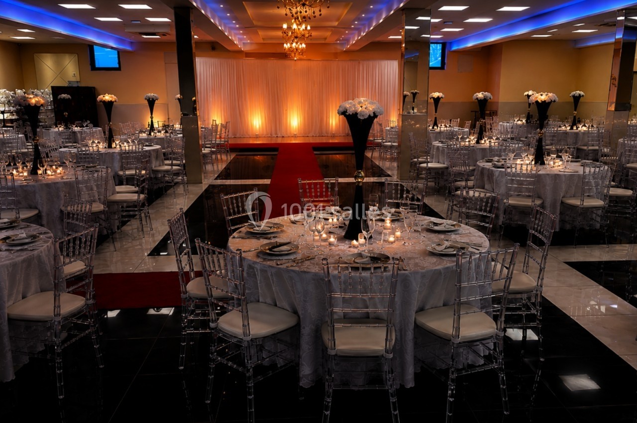 Salle de réception élégante avec tables rondes dressées, nappes grises, chaises transparentes et éclairage chaleureux.