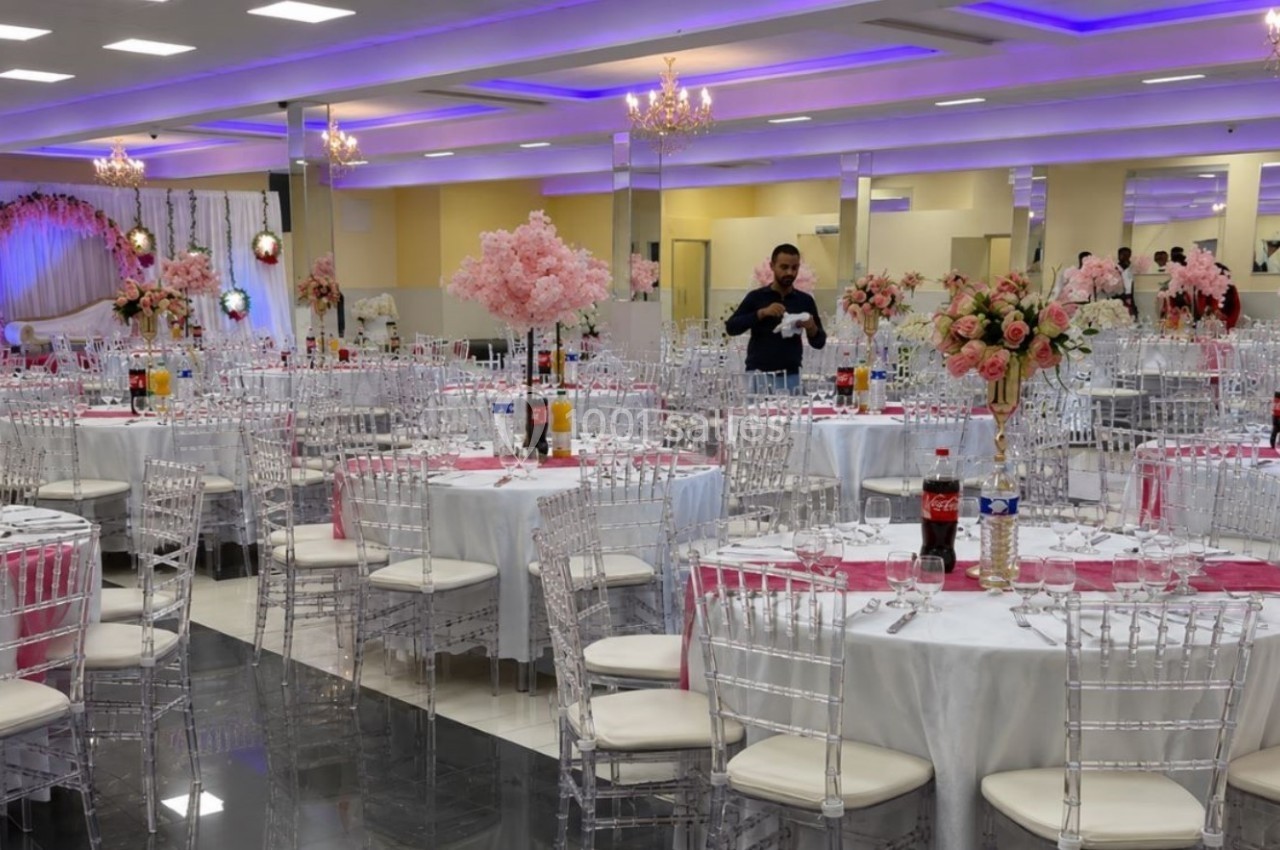 Salle de réception décorée avec des tables rondes, nappes blanches, chaises transparentes et arrangements floraux roses.