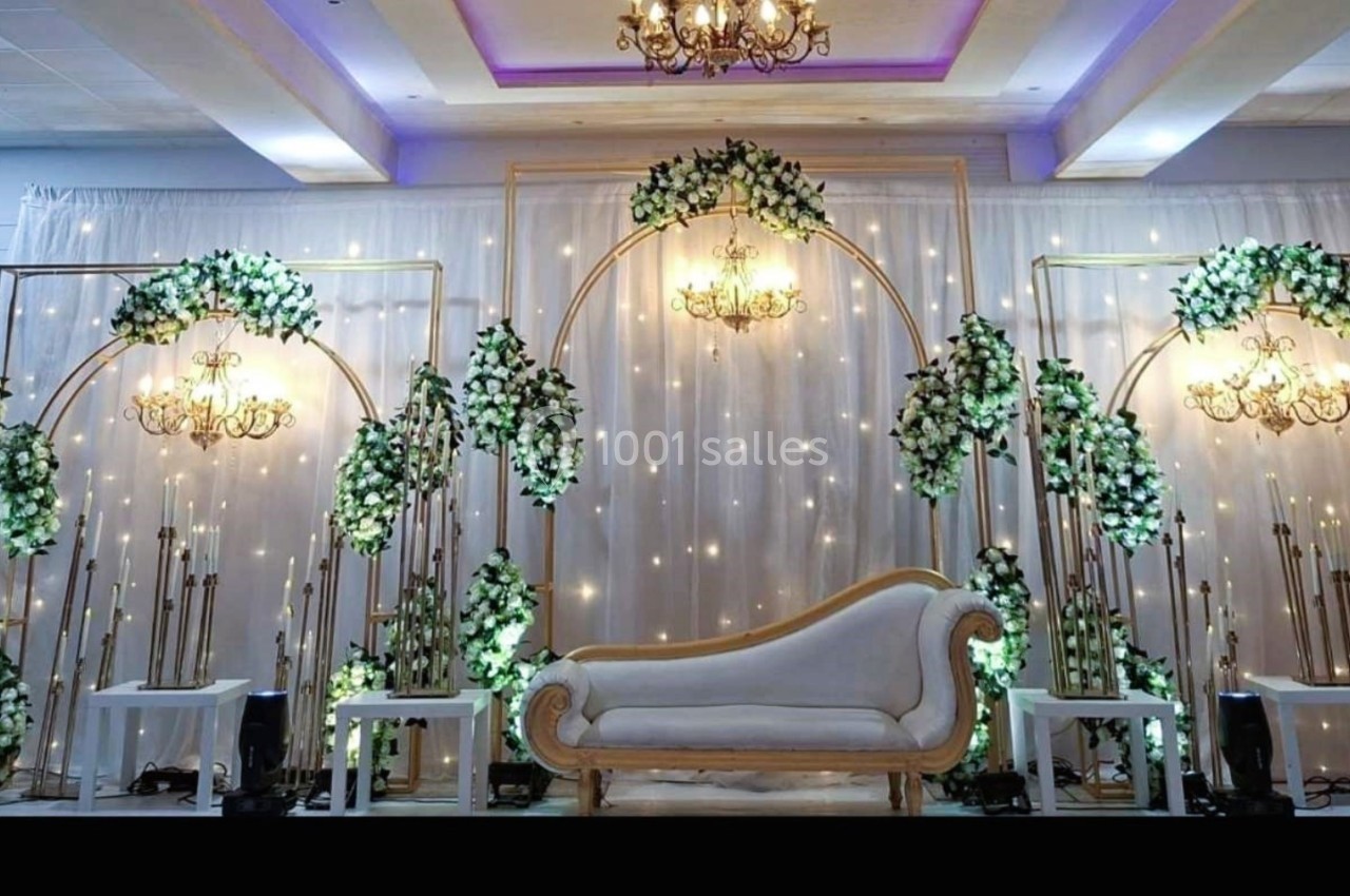 Décor de scène avec canapé beige, arches dorées ornées de fleurs blanches et éclairage chaleureux.