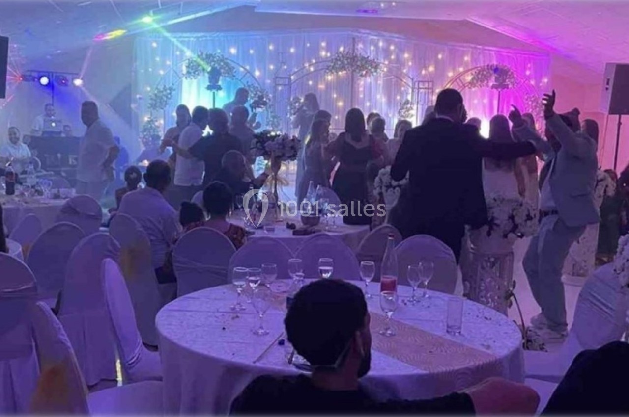 Salle de réception décorée, invités dansant et discutant autour de tables dressées, éclairage festif en arrière-plan.