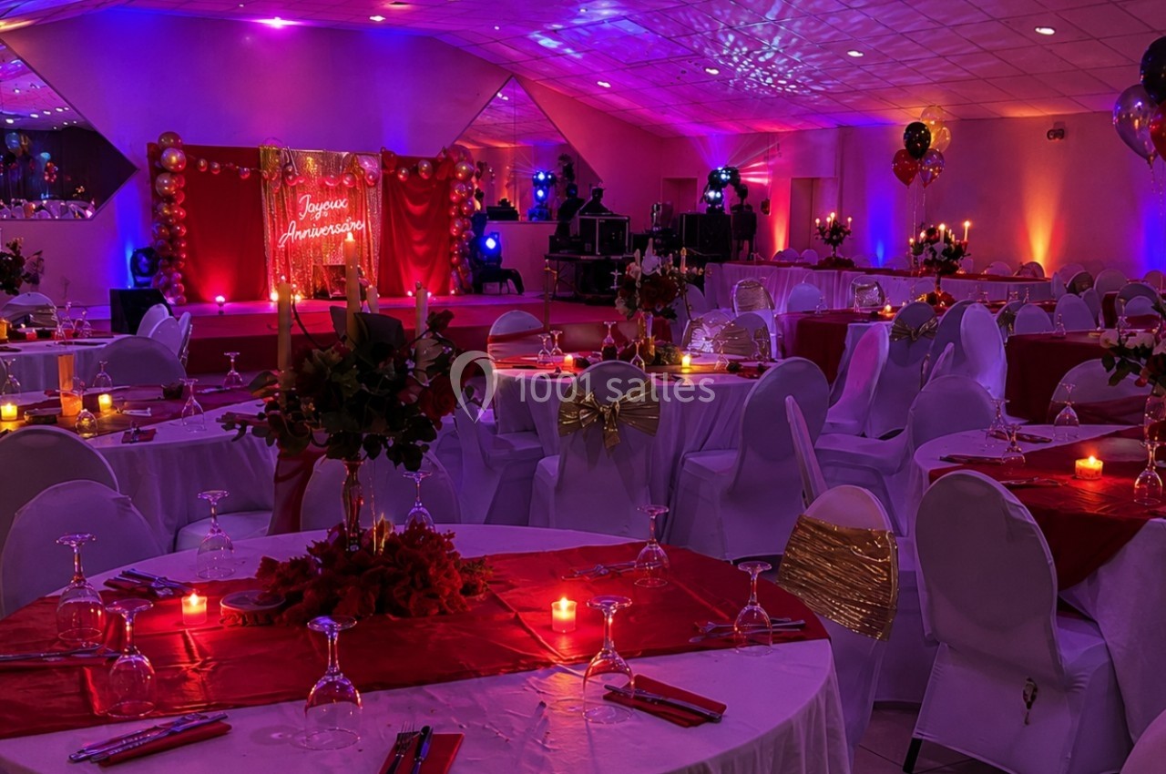 Salle de réception décorée avec des nappes rouges, bougies, ballons et éclairage tamisé pour un événement festif.