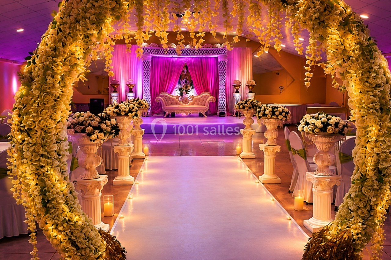 Salle de réception décorée avec des fleurs, un arc floral illuminé et une scène ornée d'un canapé élégant.