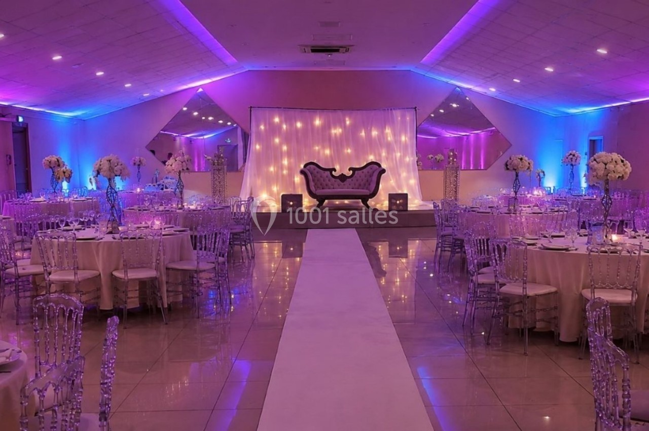 Salle de réception décorée avec des tables rondes, des chaises transparentes et un éclairage violet et bleu.
