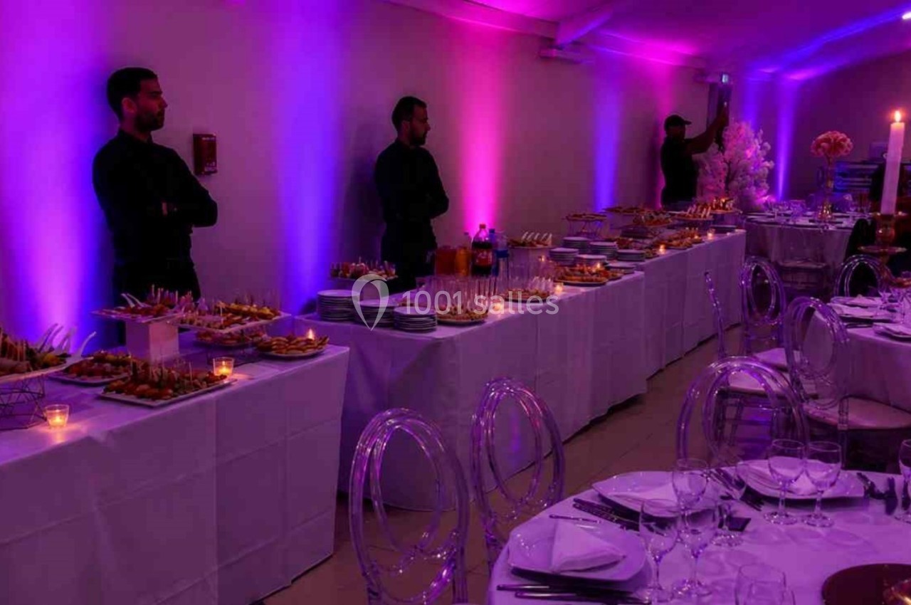 Salle de réception éclairée en violet avec tables dressées et buffet garni, surveillé par des serveurs.
