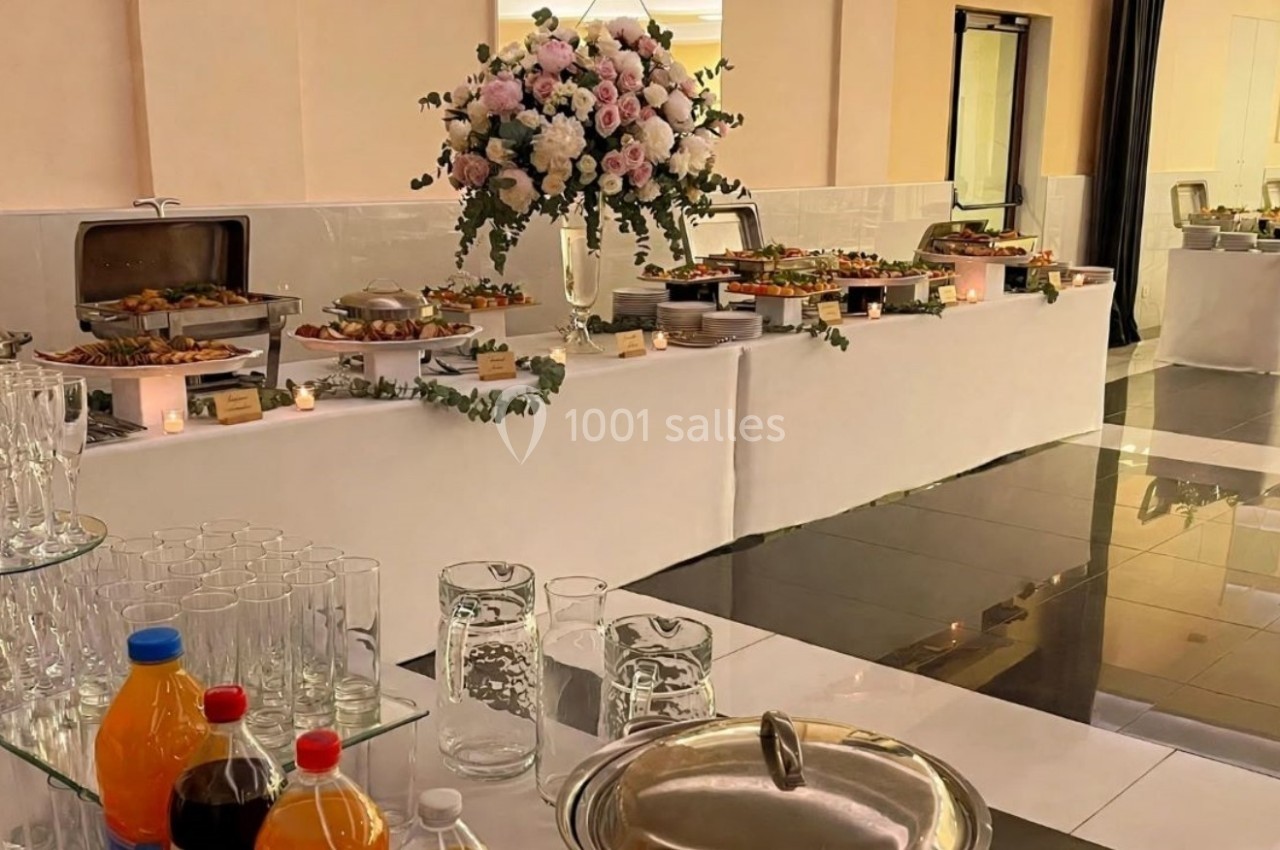 Buffet de réception avec plats variés, boissons et décoration florale dans une salle lumineuse au sol carrelé.
