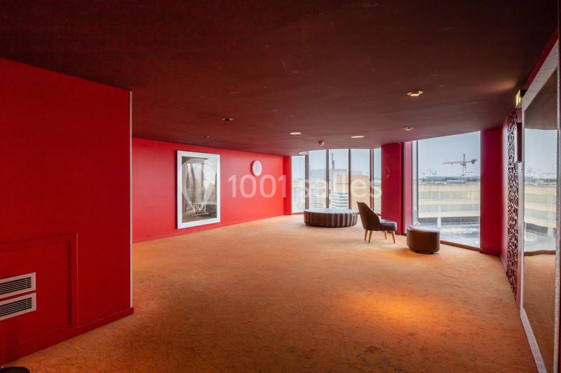 Salle lumineuse avec murs rouges, moquette orange, fauteuils près d'une baie vitrée donnant sur des bâtiments et grues.