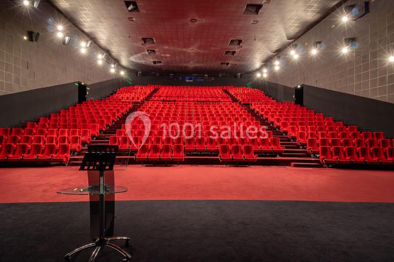 Salle de conférence vide avec des rangées de sièges rouges et un pupitre au premier plan.